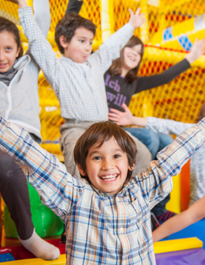 Un jeune garçon souriant les bras levés avec des autres enfants derrières lui qui lèvent aussi les bras en l'air dans la plaine de jeux couverte